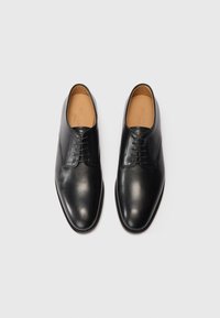 Pari di scarpe eleganti in pelle lucida nera con lacci, interni beige, posizionate affiancate su uno sfondo grigio chiaro.