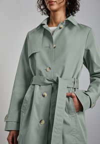 Jasnozielony trenchw coat wykonany z gładkiego materiału, z paskiem w talii, dużymi guzikami i kieszenią z klapką na piersi.