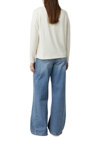 Eine Frau mit langen braunen Haaren trägt ein lockeres weißes Longsleeve-Oberteil und weit geschnittene hellblaue Jeans, steht mit dem Rücken zu einer weißen Hintergrund.