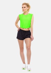 Neon zelena kratka majica i crne šortse s vezicom u struku. Model nosi bijele cipele s zelenim detaljima. Obična bijela pozadina.