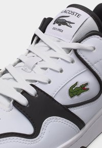 Λευκό και μαύρο αθλητικό παπούτσι Lacoste με λευκά κορδόνια, πράσινο λογότυπο κροκόδειλου στο πλάι και ετικέτα Lacoste στη γλώσσα με την επωνυμία και το έτος.