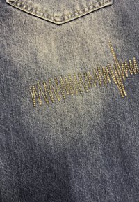 La tela de mezclilla en azul oscuro presenta un distintivo patrón de bordado dorado en zigzag cerca del dobladillo, resaltando una superficie texturizada.