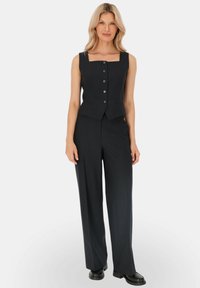 Czarna kamizelka w paski bez rękawów z kwadratowym dekoltem i przednimi guzikami, w połączeniu z szerokimi czarnymi spodniami i butami na płaskiej podeszwie.