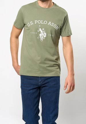 Mand iført olivengrøn U.S. Polo Assn. t-shirt med hvidt logo af to polospillere på heste og blå jeans.