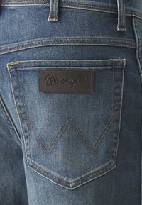 Ziloni džinsi ar aizmugures kabatu, kurai ir brūna ādas Wrangler etiķete un zigzaga šuvju raksts virs kabatas.