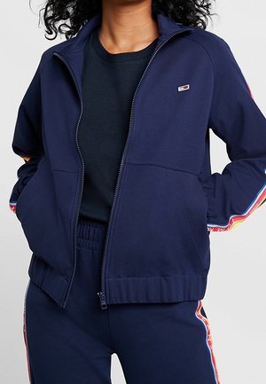 Personne portant un survêtement bleu marine avec une veste à fermeture éclair et un pantalon assorti, tous deux avec des bandes latérales colorées et un petit logo sur la poitrine.