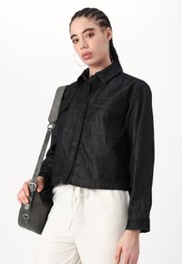 Chaqueta de mezclilla negra con cierre de botones, dos bolsillos en el pecho y mangas largas. Combinada con un bolso gris de hombro sobre un fondo liso.