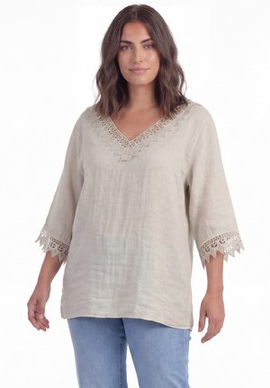 Femme portant un chemisier beige à col en V avec une bordure en dentelle au niveau du col et des manches, associée à un jean bleu clair, debout devant un fond blanc.