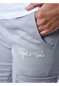 Grijze joggers met een zijzak en witte borduring met de tekst "Project x Paris." Materiaal lijkt zacht met een gladde textuur.