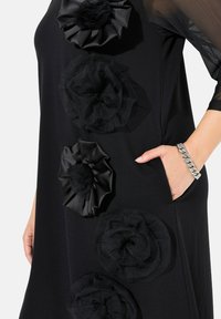 Schwarzes Kleid mit geschichteten Stoffblumen in Schwarz. Verfügt über eine Tasche, transparente Ärmel und eine glatte Textur. Silberschmuck-Akzent am Handgelenk.