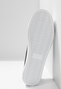 Černé tenisky s bílou gumovou podrážkou s herringbone vzorem a strukturovaným povrchem. Design je nízkoprofilový s minimálním zdobením.