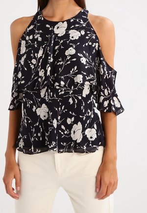 Marineblauwe cold-shoulder blouse met witte bloemenprint, ruche details en ellebooglange mouwen, gedragen met off-white broek.