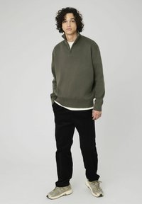 Jeune personne avec des cheveux bouclés, portant un pull zippé vert olive, un pantalon noir et des baskets beiges, se tenant devant un fond clair uni.
