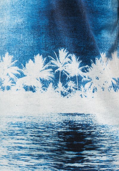 Μπλε και λευκό σχέδιο υφάσματος που απεικονίζει μια τροπική παραθαλάσσια περιοχή με φοίνικες και ήρεμες αντανακλάσεις στο νερό.