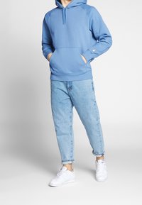Sudadera azul claro con bolsillo tipo canguro, puños acanalados y capucha con cordón, combinada con jeans azul claro y zapatillas blancas.