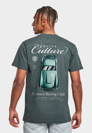 Mann med flettet hår iført mørkegrønn T-skjorte med klassisk grønn racerbilsmotiv og teksten "Motor Culture" og "Monaco Racing Club" på baksiden.
