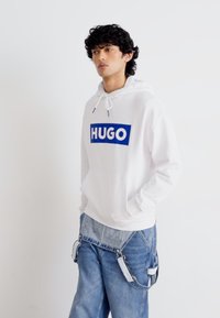 Hvit hettegenser med blå "HUGO"-logo, snorhette, kengurulomme; kombinert med løse denimjeans med unike stroppdetaljer.