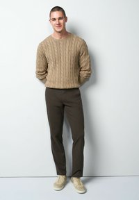 Beige kabelgebreide trui, bruine geruite broek en beige sneakers. Staande houding tegen een lichte achtergrond, handen ontspannen langs de zij.