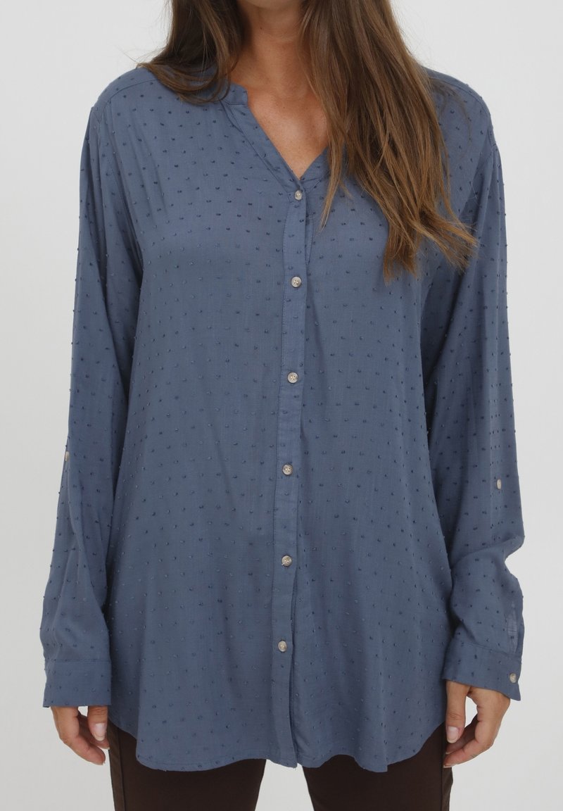 Femme portant un chemisier bleu ample avec une petite texture de petits points en relief et des boutons sur le devant, assorti à un pantalon foncé.