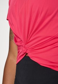 Ružová, krátkopensová blúzka s uzlom v oblasti pásu, vyrobená z elastického materiálu; v kombinácii s čiernymi nohavicami. Hladká textúra a priliehavý strih.