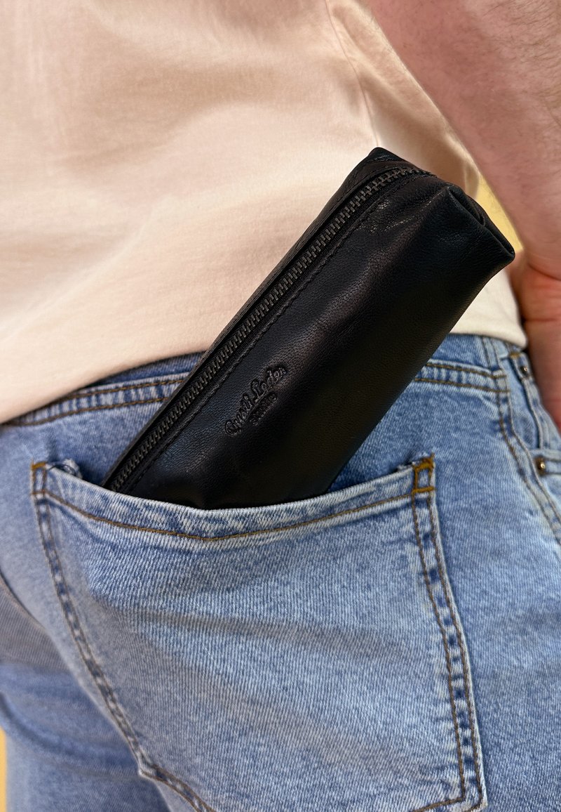 Schwarzes Leder-Mäppchen mit Reißverschluss, teilweise sichtbar in einer hellblauen Denim-Tasche. Strukturiertes Oberflächenmaterial, rechteckige Form.