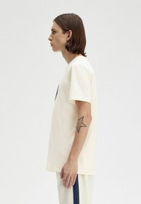 Camiseta de algodón color crema con mangas cortas, cuello redondo y un gráfico negro sencillo. El modelo tiene el pelo largo y un tatuaje visible en el brazo izquierdo.