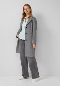 Cappotto grigio in misto lana con rever a punte, due grandi tasche frontali e bottoni neri. Abbinato a una camicia azzurra chiara e jeans grigi a gamba larga.