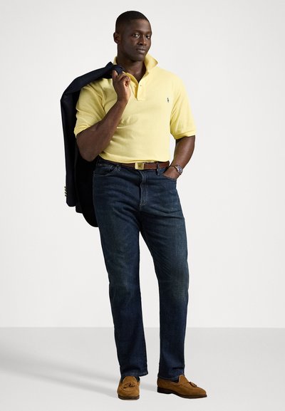 Camisa polo amarela, sapatilhas de camurça castanhas, calças de ganga azul-escuras e um blazer escuro pendurado sobre um ombro, de pé com confiança.