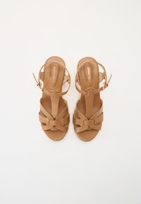Sandales synthétiques marron avec une semelle plate, présentant un design de brides entrelacées, une boucle ajustable à la cheville et une semelle intérieure texturée.