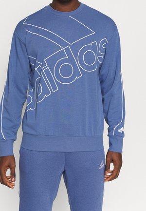 Mann trägt blauen Adidas-Sweatshirt und passende Sweatpants mit weißen Adidas-Logos und Streifen an Ärmeln und Hosen.