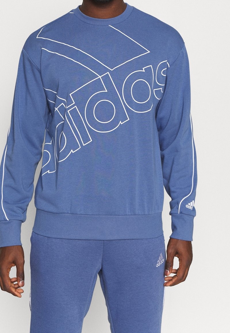 Homme portant un sweat Adidas bleu et un pantalon de survêtement assorti avec des logos blancs Adidas et des bandes sur les manches et le pantalon.