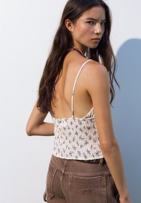 Hvid blomsteret crop top med tynde stropper og smock-design, parret med brune højtaljede bukser med et logo-detalje bagpå.