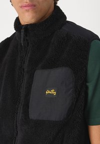 Black fleece vest featuring a high collar, zip closure, and a contrasting fabric pocket with a yellow logo accent. Textured and warm design.
