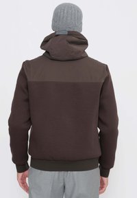 Felpa con cappuccio marrone con design bicolore; tessuto morbido e polsini elasticizzati; include cappuccio e orlo a coste, indossata sopra pantaloni grigio chiaro.