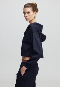 Sudadera corta de color azul marino hecha de una tela suave, con capucha ajustable, detalle de cremallera y puños de canalé. El modelo lleva pantalones oscuros a juego.