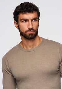 Beige pitkähihaisen neuleen, jossa on teksturoitu neulontamalli, ympärillä pyöreä pääntie ja istuva siluetti, malli, jolla on lyhyet tummat hiukset.