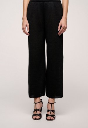 Underkrop iført løstsiddende sorte bukser og sorte højhælede sandaler med stropper, stående mod en ensfarvet lys baggrund.