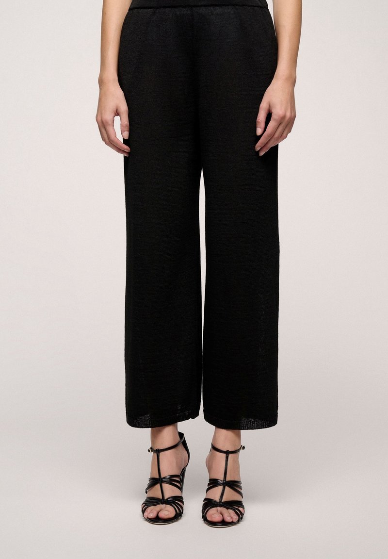 Partie inférieure du corps vêtue d’un pantalon noir ample et de sandales à talons hauts noires à brides, debout devant un fond clair uni.