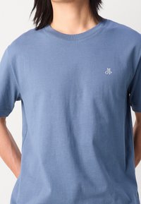 Modré bavlněné tričko s krátkým rukávem, na levé hrudi s malým bílým logem. Hladká textura a klasický kulatý výstřih.