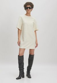 Crème gekleurde jurk met korte mouwen en een relaxte pasvorm, gecombineerd met kniehoge zwarte leren laarzen en oversized zwarte zonnebril.