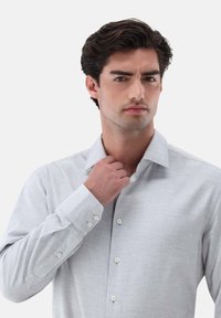 Camicia a maniche lunghe grigia chiara con tessuto texturizzato e colletto. Presenta bottoni bianchi e una vestibilità rilassata. Uomo che tocca il colletto.