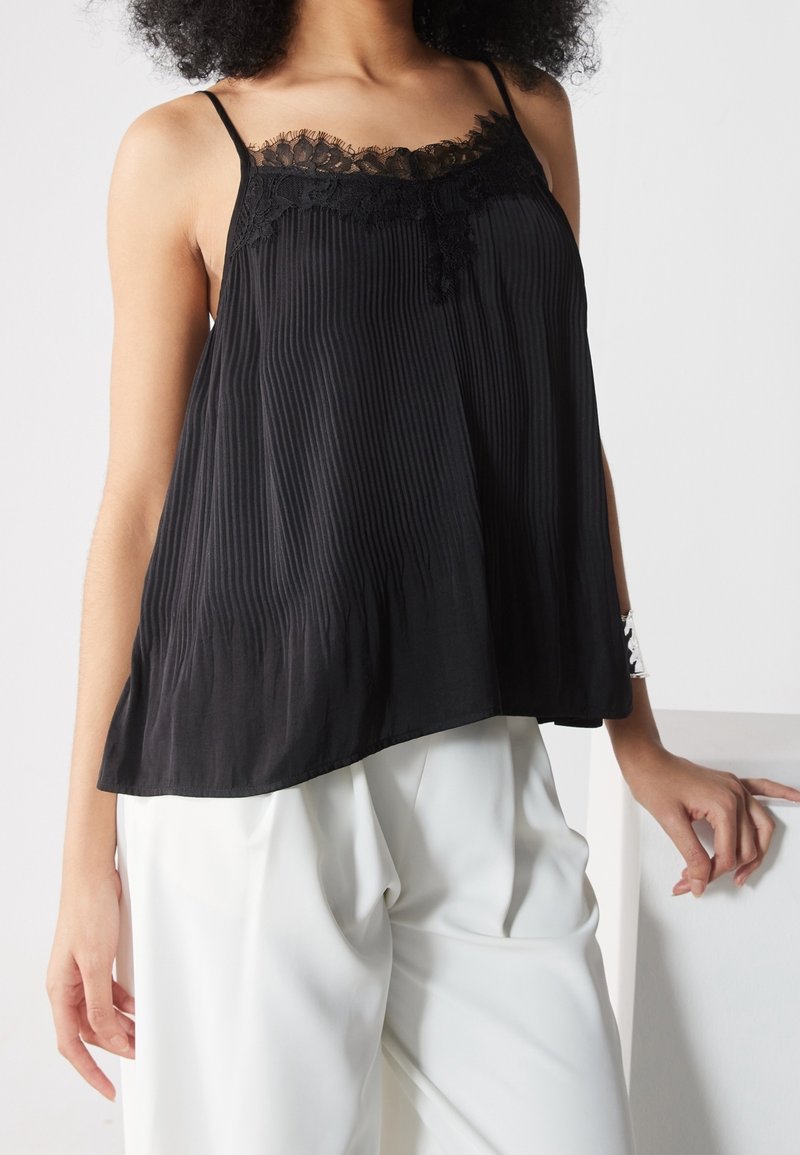Femme portant un haut plissé noir sans manches avec bordure en dentelle et un pantalon blanc ample, appuyée sur une surface blanche avec la main gauche.