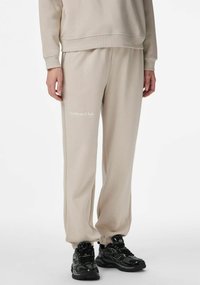 Beżowe spodnie dresowe ze ściągaczami przy nogawkach, miękka tkanina i nadrukowane logo „Aesthetic Club” na lewej udzie. Do tego czarne sportowe buty.