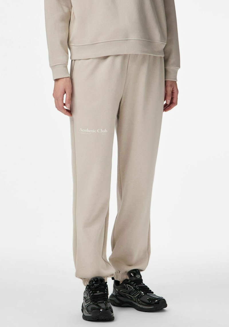 Beżowe spodnie dresowe ze ściągaczami przy nogawkach, miękka tkanina i nadrukowane logo „Aesthetic Club” na lewej udzie. Do tego czarne sportowe buty.