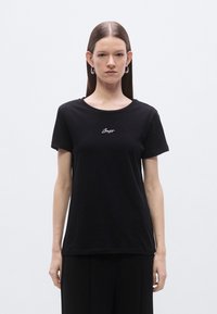 Črna bombažna majica z okroglim izrezom in kratkimi rokavi, z belim vezenim napisom na prsih. Gladek tekstura, sproščen kroj.