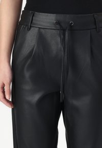 Vrouw in zwarte neplederen broek met een trekkoord in de taille en riemlussen, weergegeven van halverwege het bovenlijf tot halverwege de bovenbenen.