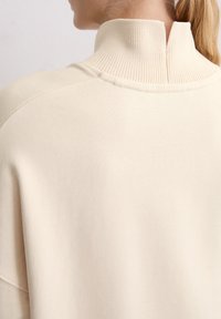 Beżowy sweter z wysokim, żeberkowym kołnierzem i rozcięciem z tyłu. Miękka, teksturowana tkanina z gładkimi szwami i opadającymi szwami ramion.