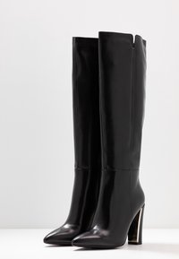 Svarta knähöga stövlar i slät läder med spetsig tå och en chunky metallklack för ökad designkontrast.