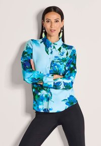 Woman with long dark hair wearing blue floral button-up blouse and black pants, standing with arms crossed against a plain background.