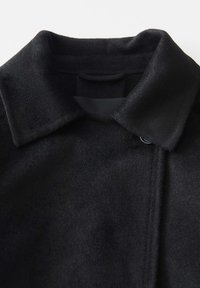 Cappotto di lana nera con colletto, caratterizzato da una texture morbida e una chiusura con un singolo bottone. Il tessuto appare liscio e spesso.